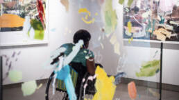 Gallery attendee in wheelchair photographed through paint splattered window admiring artwork in art gallery.