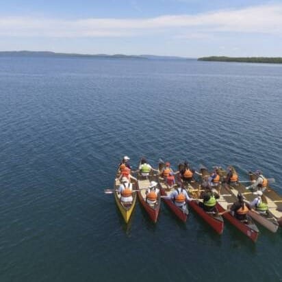 6 canoes right next to each other on a large lake. 2 pople are holding paddles in each canoe