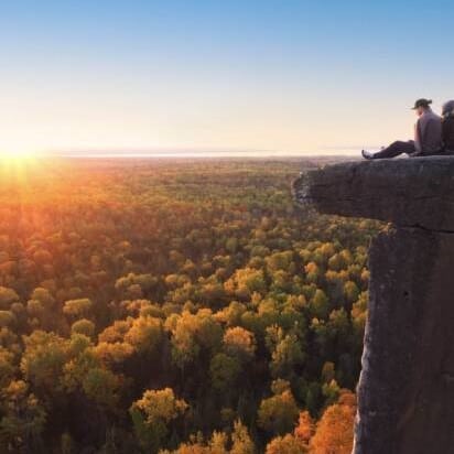 A person sitting on a rock structure very high up, overlooking thousands of trees with the sun setting in the distance