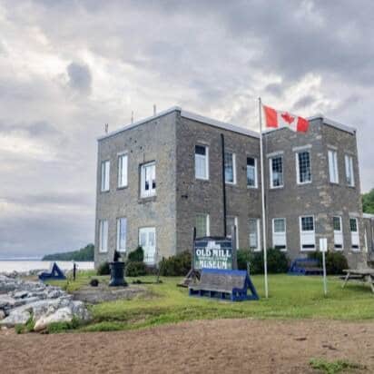 A large stone building next to a lake. A Canadian flag is next to the building and a sign that reads "Old Mill"
